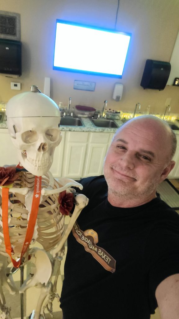 Brandon, co-owner of Fancy’s Day Spa and Texas Massage Academy, smiling and posing with a classroom skeleton model inside a massage therapy training room, wearing a Texas Massage Academy T-shirt, showcasing a fun and welcoming atmosphere.