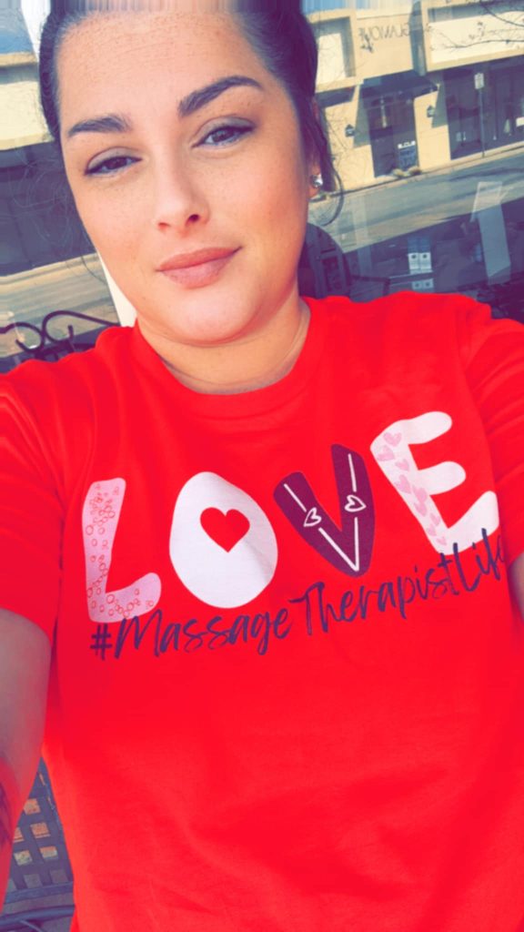 Brittney, night instructor and spa therapist at Fancy’s Day Spa, smiling while seated in front of Fancy’s storefront, wearing a bright red T-shirt with the words ‘LOVE #MassageTherapistLife,’ radiating warmth and pride in her massage therapy career.