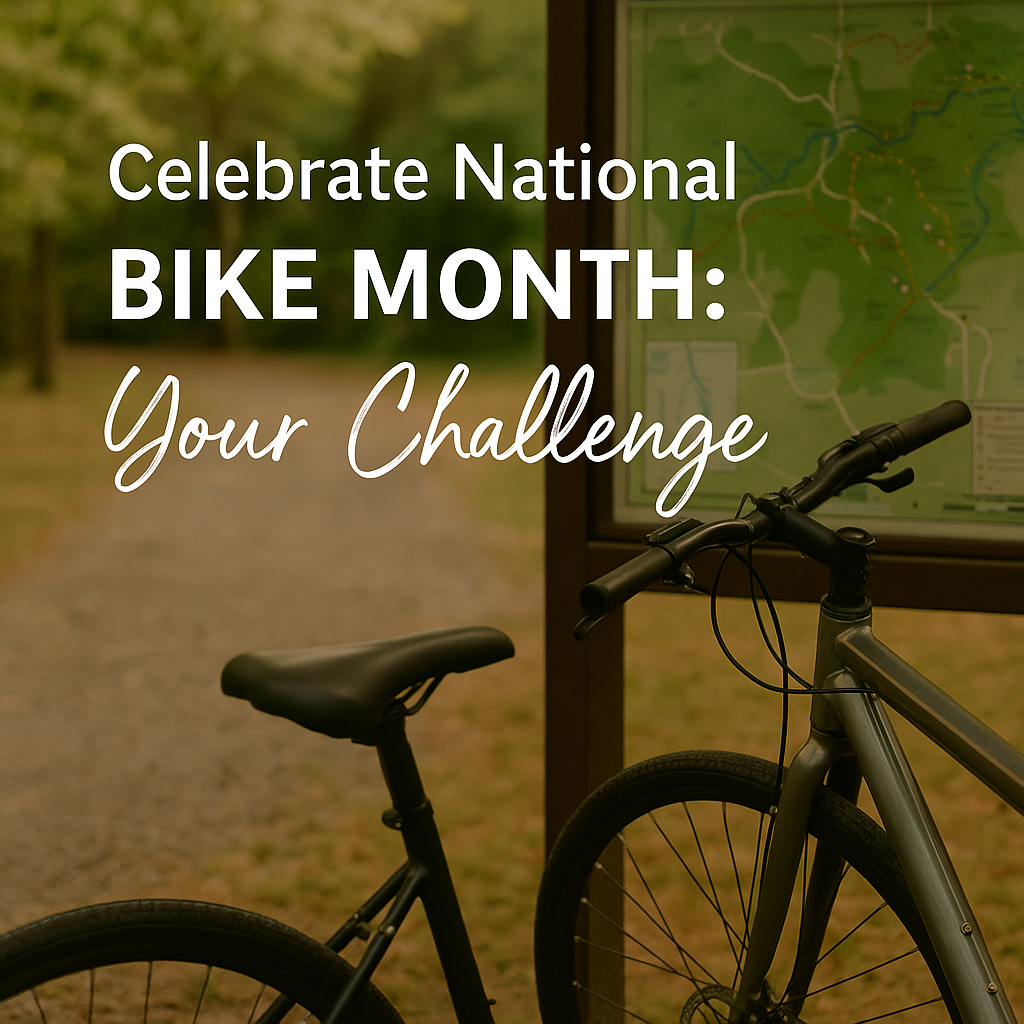 A bicycle parked next to a trail map at the entrance of a forested path, with overlaid text that reads ‘Celebrate National Bike Month: Your Challenge,’ surrounded by trees and sunlight.