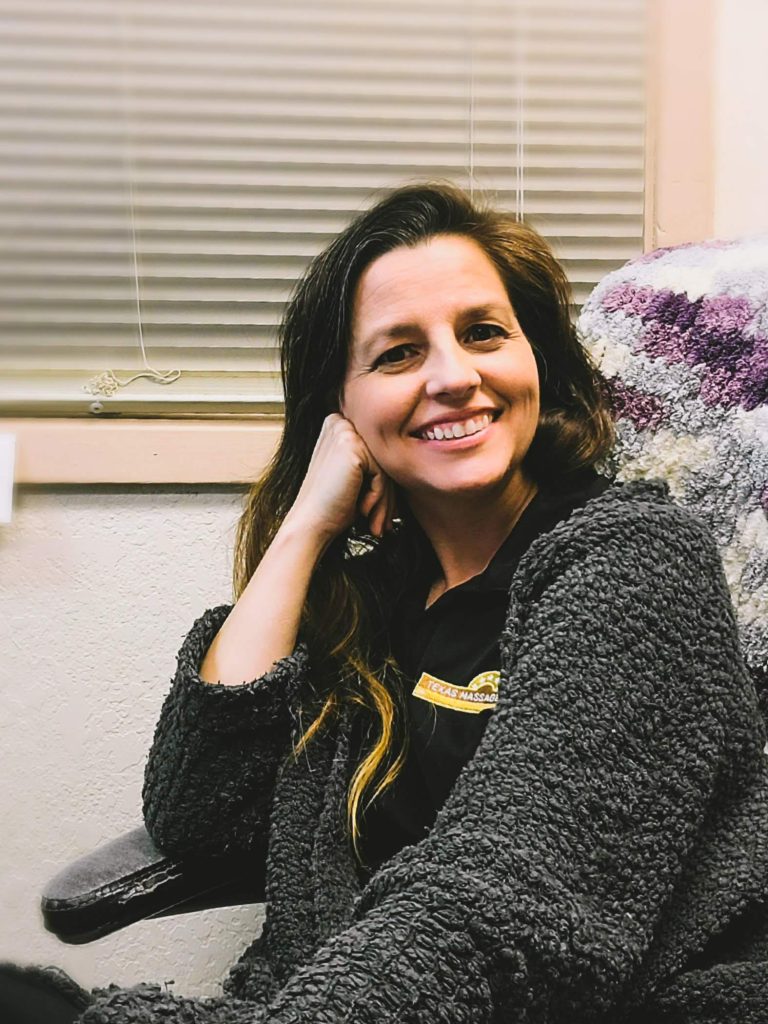 Kathleen, owner of Fancy’s Day Spa and Texas Massage Academy, smiling warmly while seated in a cozy office chair, wearing a textured gray sweater and black shirt with the spa logo, embodying a welcoming and caring presence.