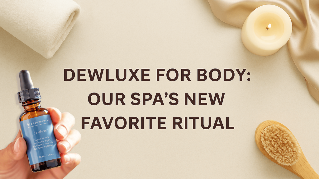 Flat lay of spa essentials including a rolled towel, candle, satin robe, and dry brush, with a hand holding a bottle of Bioelements Dewluxe for Body oil next to the headline: 'Dewluxe for Body: Our Spa’s New Favorite Ritual.