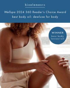 Woman in neutral-toned lounge wear applying Dewluxe to her leg, with text reading: 'WellSpa 2024 Reader’s Choice Award – Best Body Oil: Dewluxe for Body.'