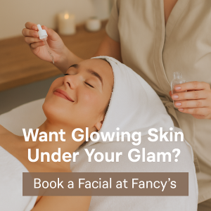 A smiling woman relaxes with eyes closed while an esthetician applies serum to her face during a facial. The spa room is softly lit with candles in the background. Text reads “Want Glowing Skin Under Your Glam? Book a Facial at Fancy’s.”
